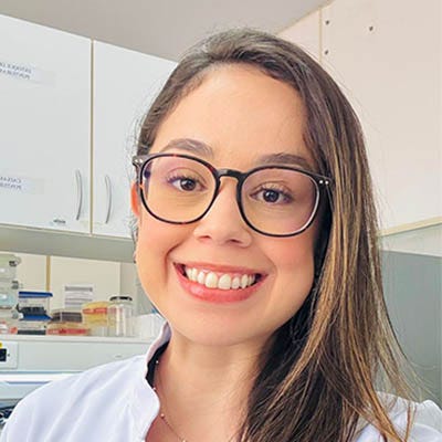 A foto mostra uma mulher jovem, de jaleco branco, sorrindo em um ambiente de laboratório, com equipamentos científicos ao fundo.