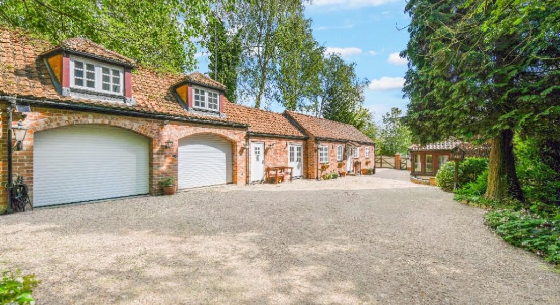 A large cottage with a brick exterior, a gravel driveway, and greenery around it A large cottage with a brick exterior, a gravel driveway, and greenery around it