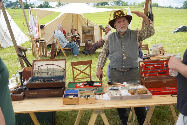 A man in Civil War attire sits in front of a white tent and a man in a gray vest of period attire stands behind a table of medical equipment.