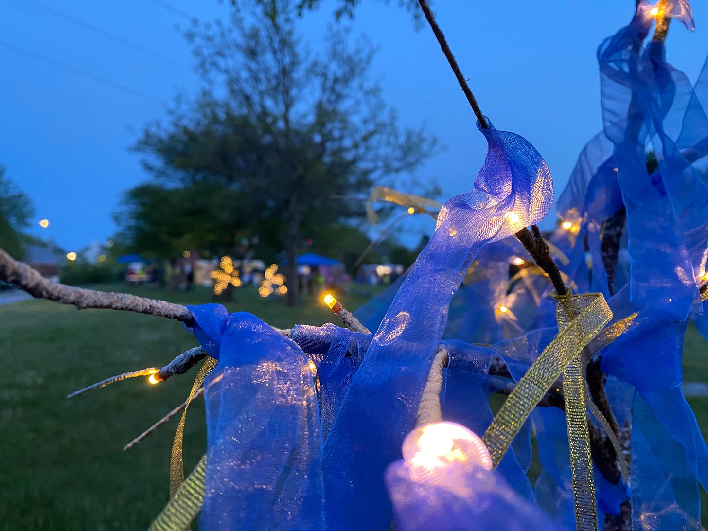 Closeup photograph of a lighted artificial tree with blue ribbons tied in its branches. Other trees and a party are visible in the background.