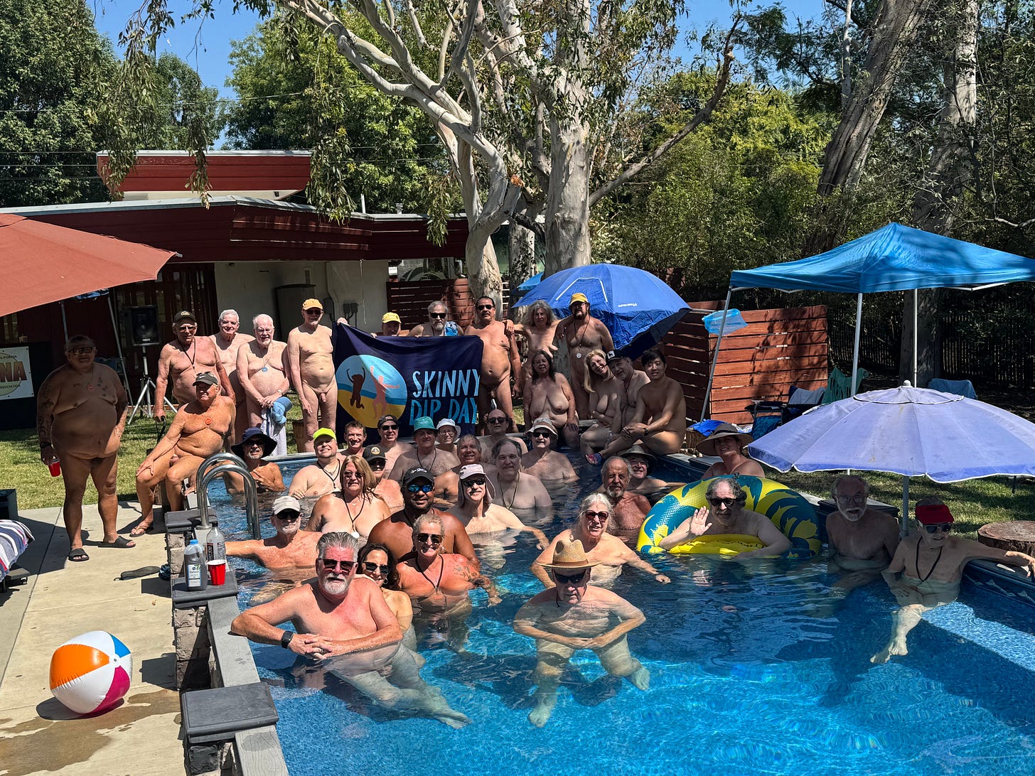 Dozens of nude people gather around and inside a backyard swimming pool under umbrellas and trees. Some are seated in the water, others pose on the deck behind a Skinny Dip Day banner. A beach ball and floaties add to the festive scene. Dozens of nude people gather around and inside a backyard swimming pool under umbrellas and trees. Some are seated in the water, others pose on the deck behind a Skinny Dip Day banner. A beach ball and floaties add to the festive scene.