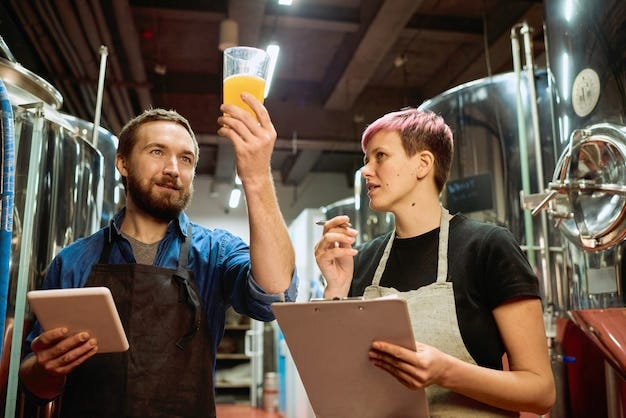 Joven cervecero barbudo en delantal mirando cerveza en vaso mientras está  de pie junto a su colega con documento durante el trabajo en equipo | Foto  Premium