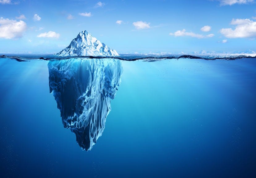 Photo of an floating iceberg, taken at ocean surface level, with the small white tip above the surface against the sky and the majority of it visible below the surface in the deep blue
