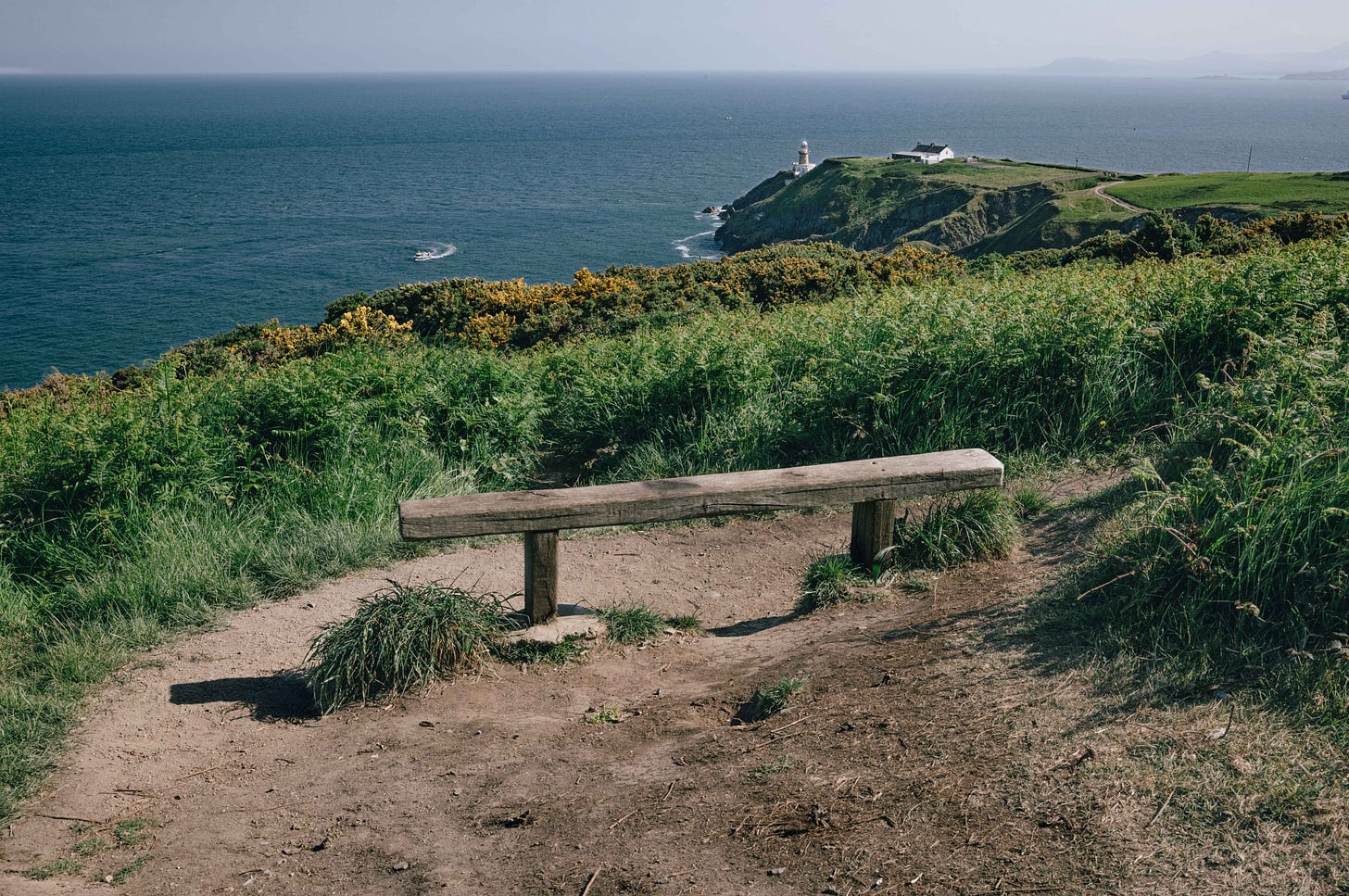 A weathered wooden bench overlooks the cliffs at Howth Head, with the Baily Lighthouse and the Irish Sea stretching into the distance. A weathered wooden bench overlooks the cliffs at Howth Head, with the Baily Lighthouse and the Irish Sea stretching into the distance.
