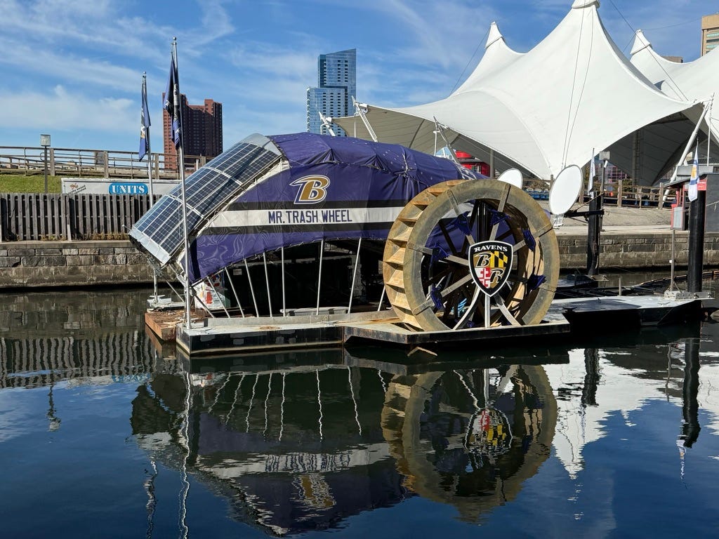 A small barge-like boat with riverboat wheels and solar panels with the name MR. TRASH WHEEL on the side and Baltimore Ravens logo and colors draped over it
