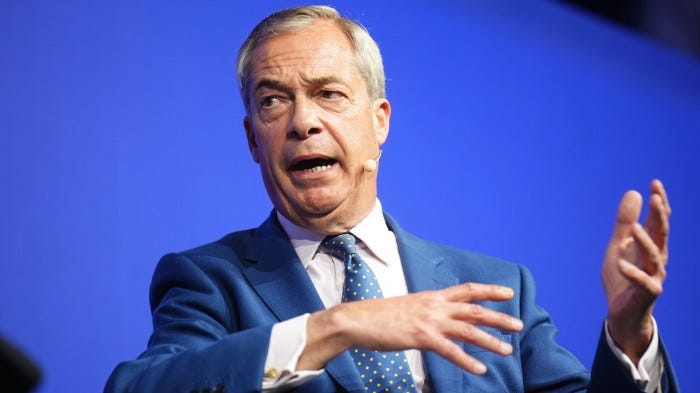 Nigel Farage speaks on stage with a microphone headset, gesturing with his hands against a blue background.