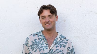 Adam wears a white and blue floral shirt and a chain. He has wavy hair