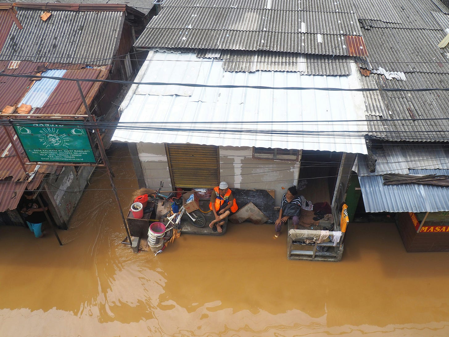An aerial photo shows a man and a woman each sitting on a chair on the porch of a house in a densely populated neighborhood that was flooded