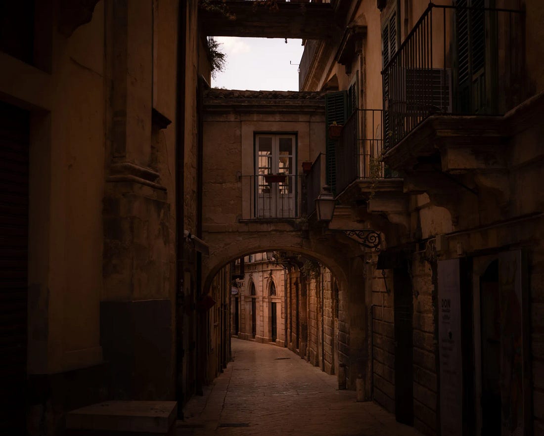 A stone arch beneath a covered passage in Modica, a narrow limestone lane running on into shadow past iron balconies. A stone arch beneath a covered passage in Modica, a narrow limestone lane running on into shadow past iron balconies.