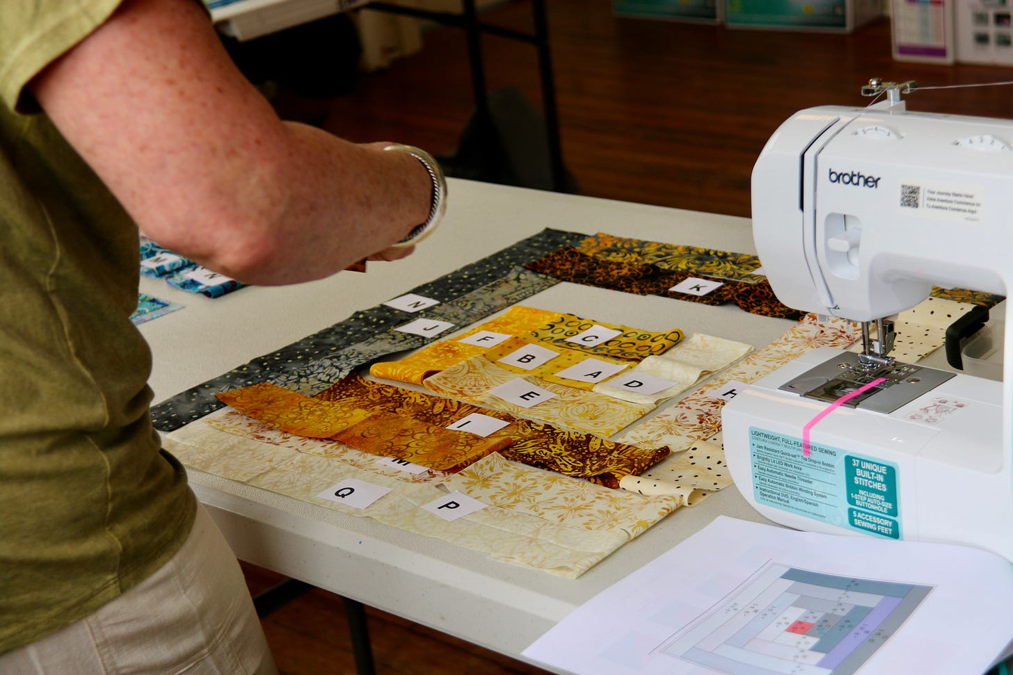 A person at a table with fabric pieces labeled with letters, preparing to sew with a Brother sewing machine.