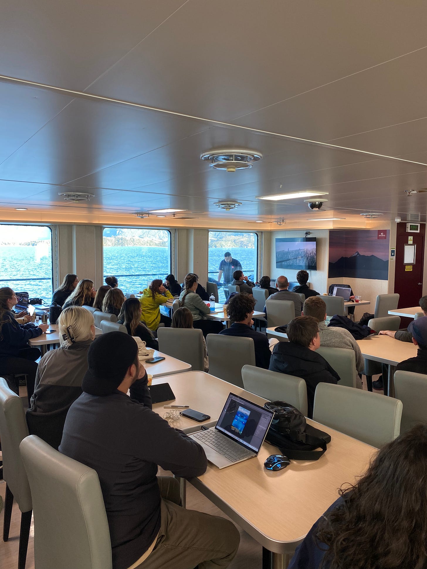 Students sitting at tables on the ferry watching a film on the TV.