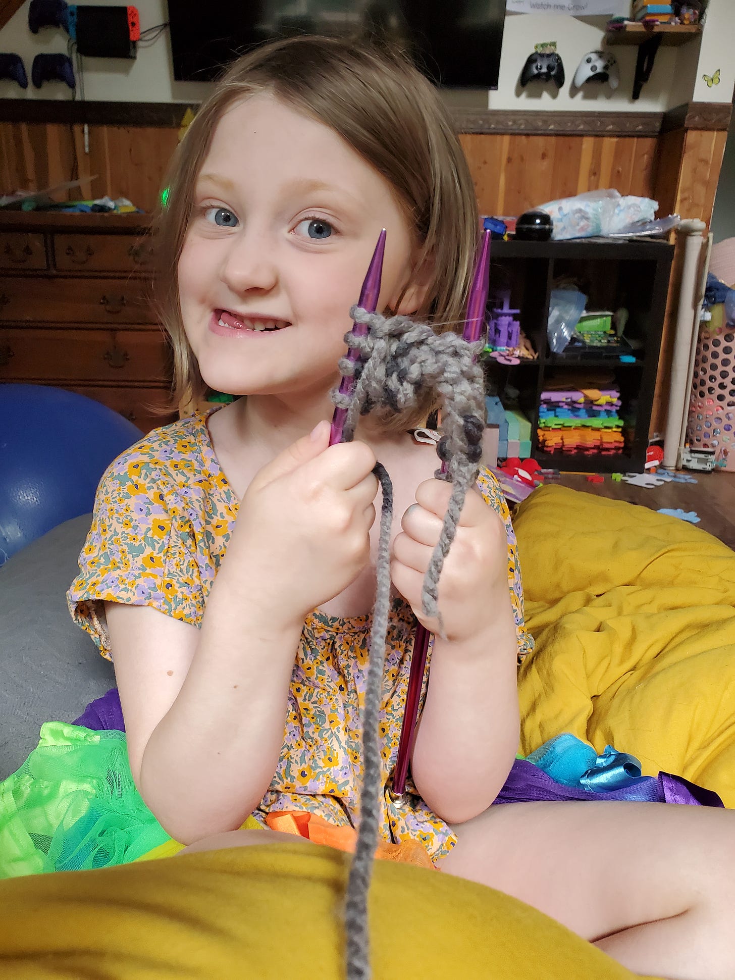 A 6-year-old smiles at the camera with her mouth quirked while proudly holding up two knitting needles with several rows of beautiful stitching.