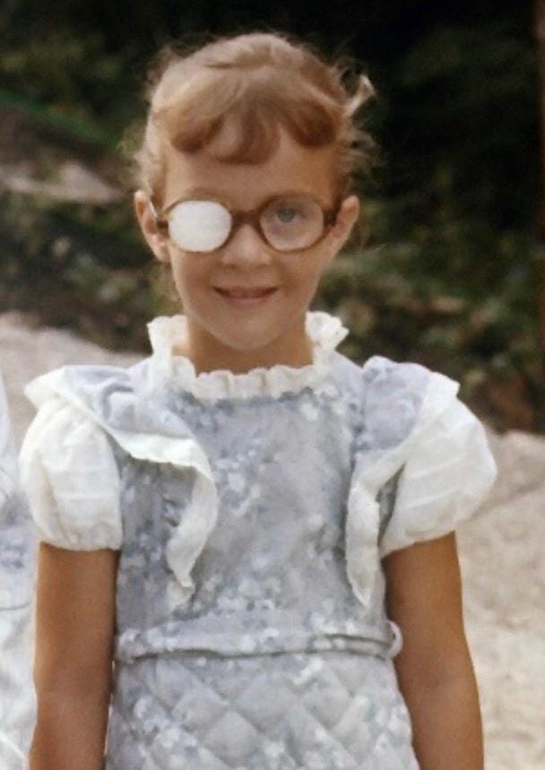 Smiling Kelly at 5 years old with a frilly blue dress, glasses and an eyepatch. 