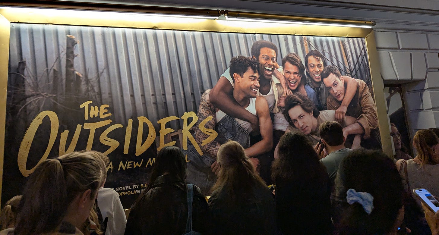 A crowd of people stands in front of a theater poster for “The Outsiders: A New Musical,” featuring six smiling cast members embracing against a corrugated metal backdrop.