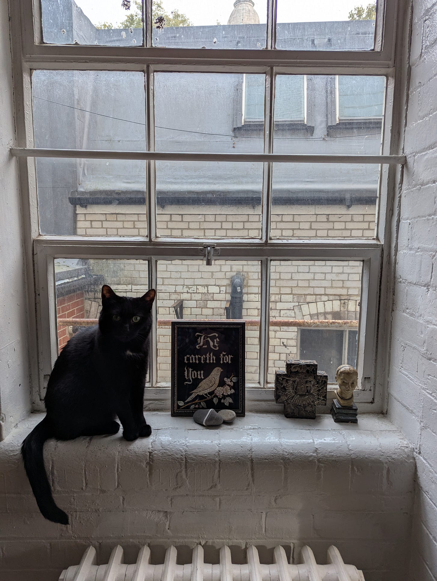 A black cat sits on a white-painted windowsill, looking directly at the camera. Behind the cat, through old sash windows, grey London rooftops and chimneys are visible. On the windowsill beside the cat are arranged: a framed embroidery reading 'he careth for you' with a bird motif, a smooth grey stone, an ornate carved wooden fragment, and a small classical bust. A white radiator is visible below the windowsill.