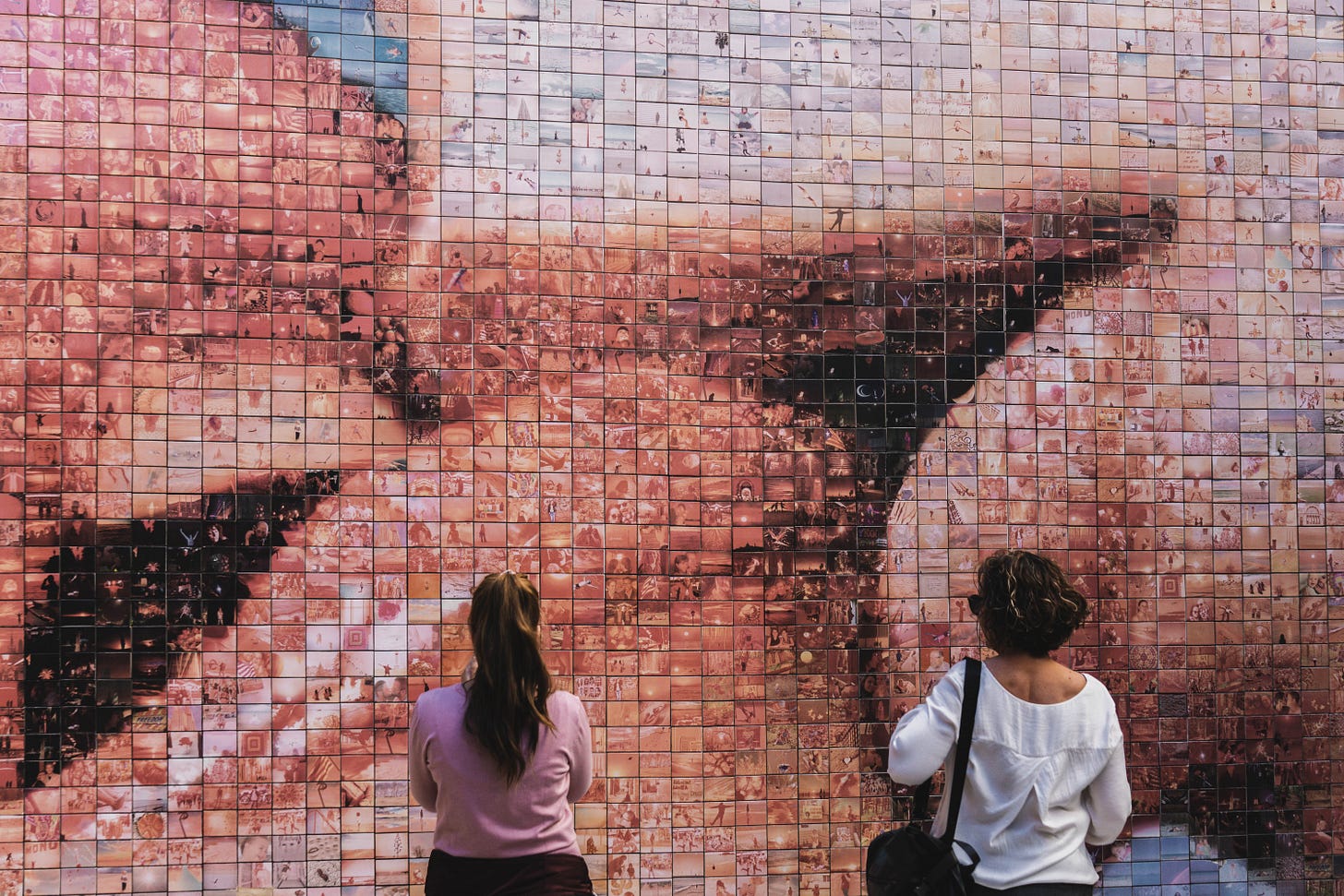 Two women are looking at street art of a mosaic of photos forming two people kissing.