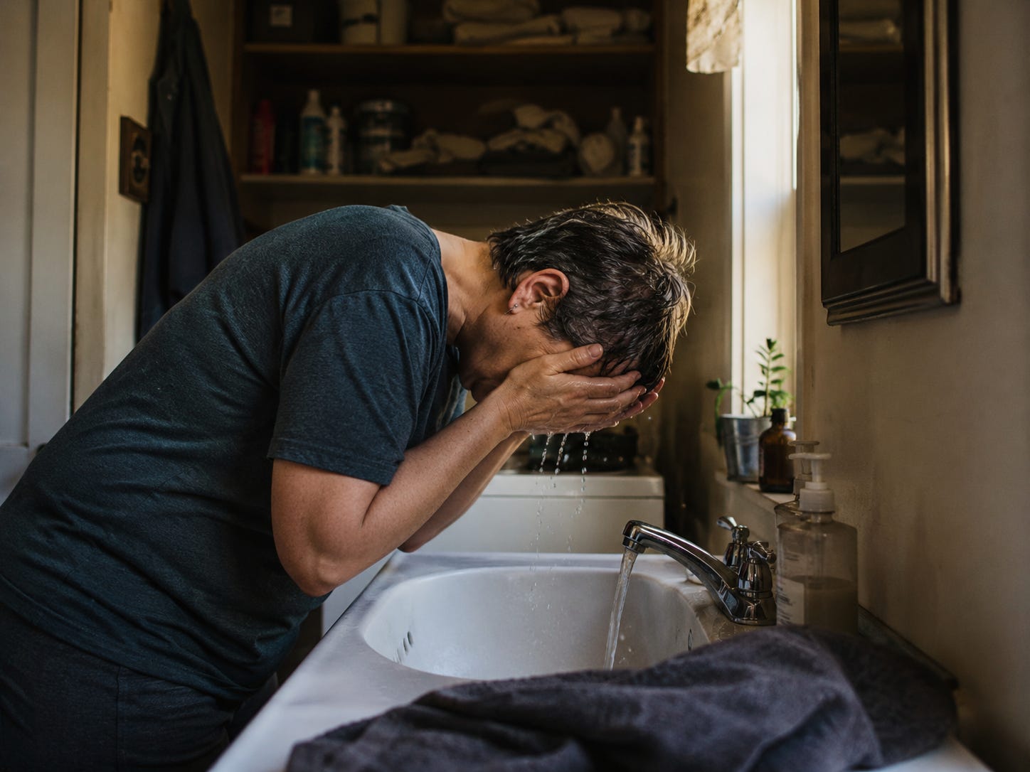 A weary woman with short wet hair leans over a bathroom sink, washing her face in soft morning light, suggesting exhaustion, adaptation, and the quiet weight of daily responsibility.