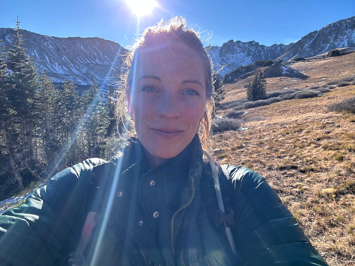 Woman hiking with the mountains and sun behind her.