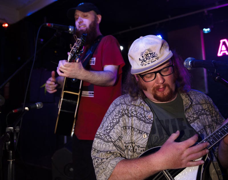 Kez, left, of Beardsnax, with a dark beard and cap at the mic with a guitar in a red T-shirt; and at right, Cai, also in hat and bearded, but with longer hair and glasses, rocking out on the mandolin, also at a mic.