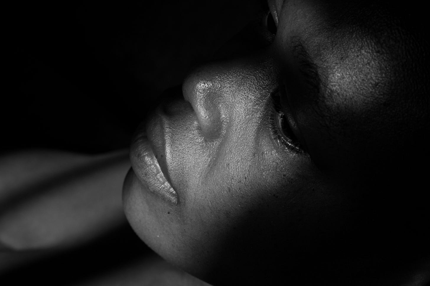 A black and white close-up photograph showing the profile of a person's face in dramatic lighting. The subject appears to be looking upward with their eyes closed, captured from below the chin and jawline. The lighting creates strong contrasts between light and shadow, emphasizing the contours of their face and neck. The image has an intimate, contemplative quality with the subject appearing peaceful or in a moment of quiet reflection.
