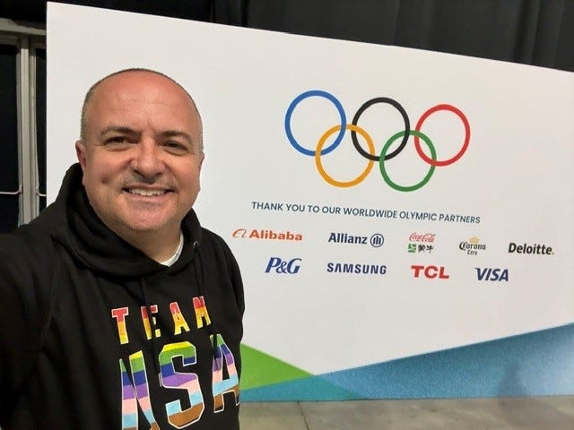 Me in Milan at the 2026 Olympic Winter Games in front of a picture with the Olympic Rings and logos of worldwide Olympic Partners.