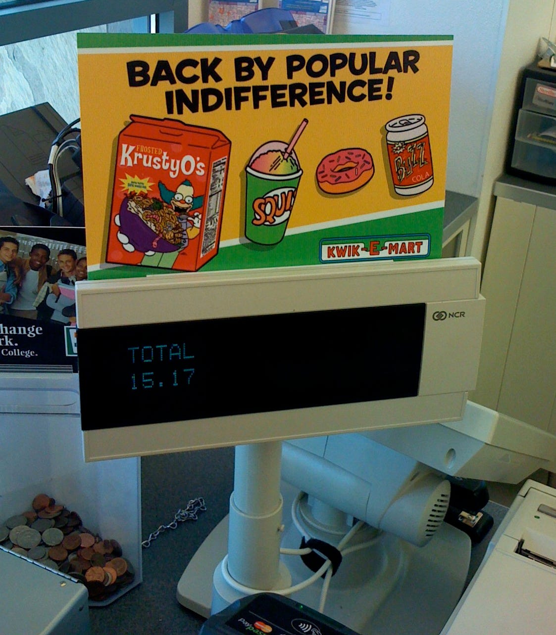 Cash register from customer's point of view. A sign above the display reads "Back by popular indifference!" and shows images of a box of Krusty-Os, a Squishee, a pink donut, and a can of Buzz Cola. There is a Kwik-E-Mart logo at the bottom of the sign.