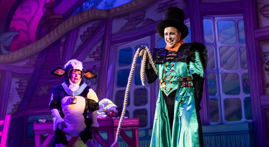 Two people on stage - a woman wearing a cow costume with a bell and a man wearing a black top hat and green coat holding a rope