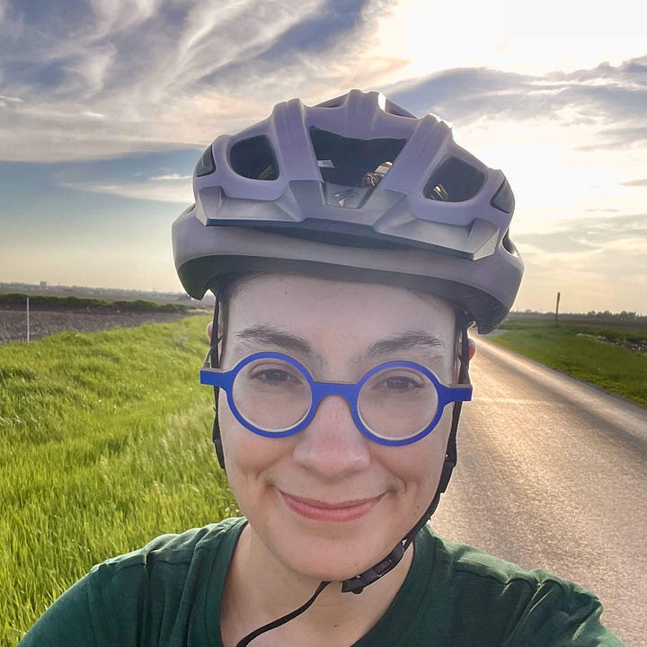 1 - A paved trail through dense foliage; 2 - A gloved hand holding a small sketchbook with water color palette clipped to it , with a landscape sketch of early spring fields; 3 - Shell wearing a bike helmet and looking at the camera; 4 - A bouquet of flowers in the water bottle holder on a bike