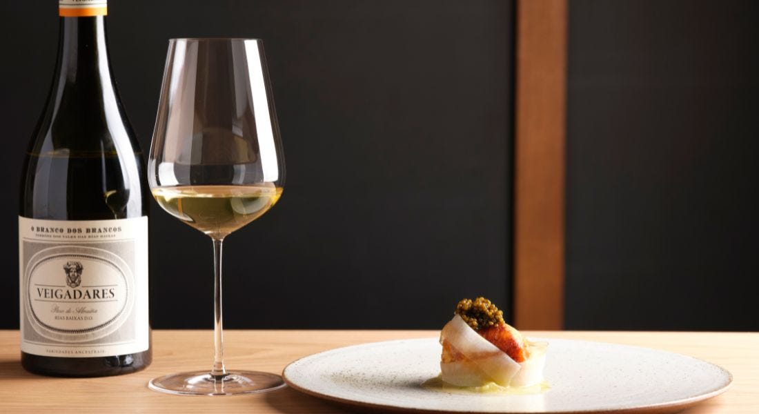 A bottle of wine, a glass of white wine, and a white plate with a small dish of food on it, with a black background behind them A bottle of wine, a glass of white wine, and a white plate with a small dish of food on it, with a black background behind them
