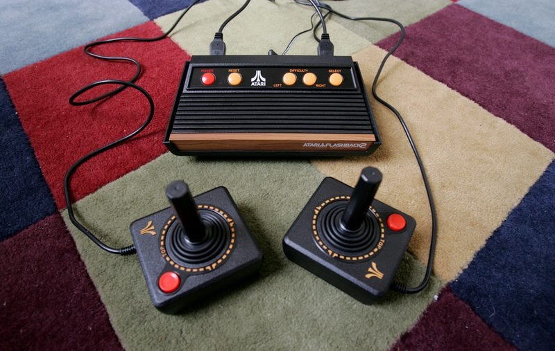 een foto van een atari 2600 spelcomputer van bovenaf gefotografeerd op een kleurrijk vloerkleed. In de voorgrond zie je twee joysticks