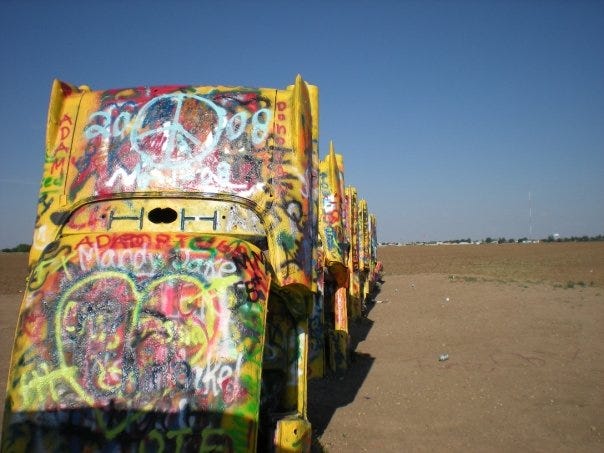 A photograph of a row of vintage Cadillacs, all painted yellow and covered in a lot of multicolored graffiti, in a flat, sandy landscape against a bright blue sky. The photo has the saturated and slightly dreamy look of an early 2000's digital camera. The car in the forground has a large white peace sign and green and yellow heart spraypainted on it.