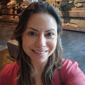 Mulher sorri para a câmera em selfie tirada em ambiente interno. Ela tem cabelos castanhos com mechas claras e veste blusa rosa, com decoração em pedra desfocada ao fundo Mulher sorri para a câmera em selfie tirada em ambiente interno. Ela tem cabelos castanhos com mechas claras e veste blusa rosa, com decoração em pedra desfocada ao fundo