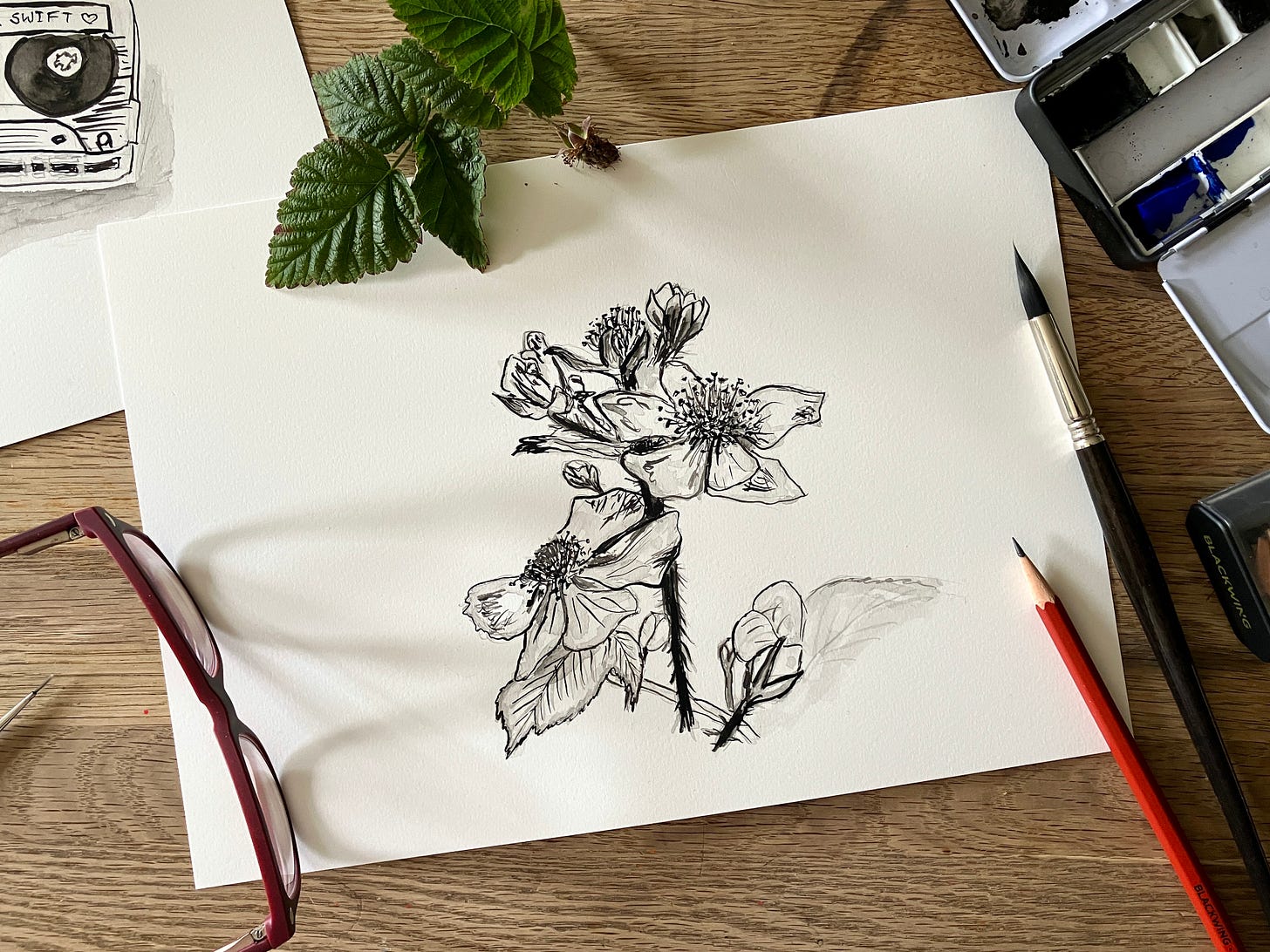 Painting of a bramble / blackberry sits atop a table with foliage and art materials surrounding it