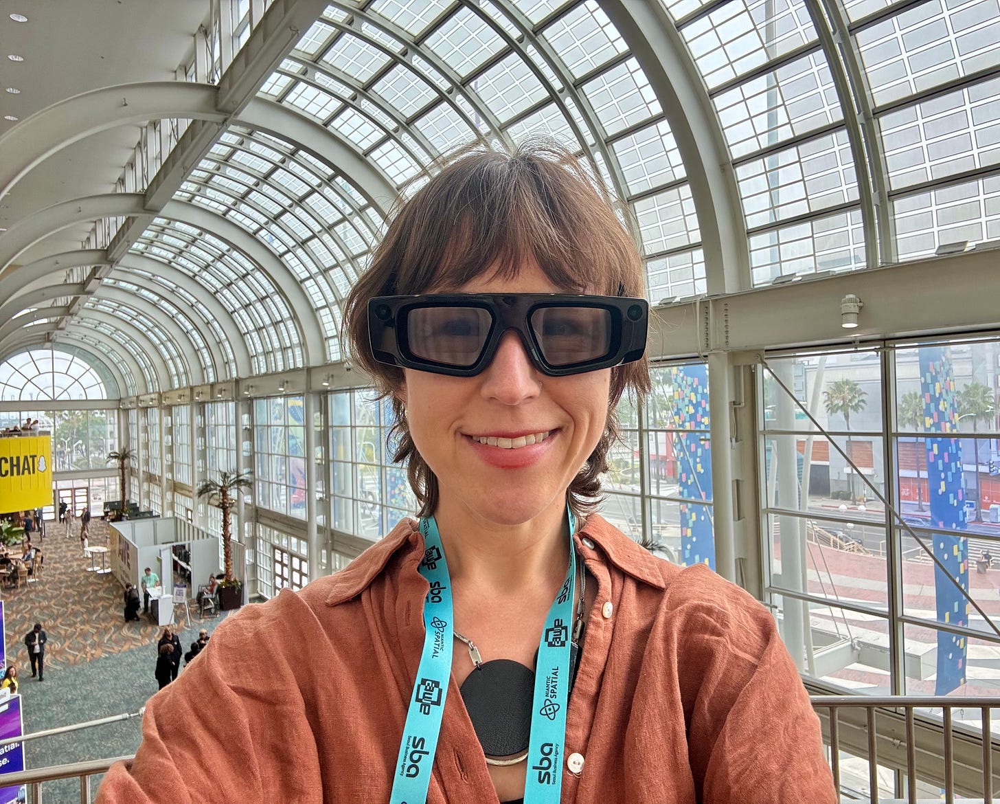 A woman wearing large black augmented reality (AR) glasses smiles for a selfie inside a convention center with a high, arched glass ceiling. She has a conference badge around her neck, and the Snapchat logo is visible on a yellow sign in the background A woman wearing large black augmented reality (AR) glasses smiles for a selfie inside a convention center with a high, arched glass ceiling. She has a conference badge around her neck, and the Snapchat logo is visible on a yellow sign in the background