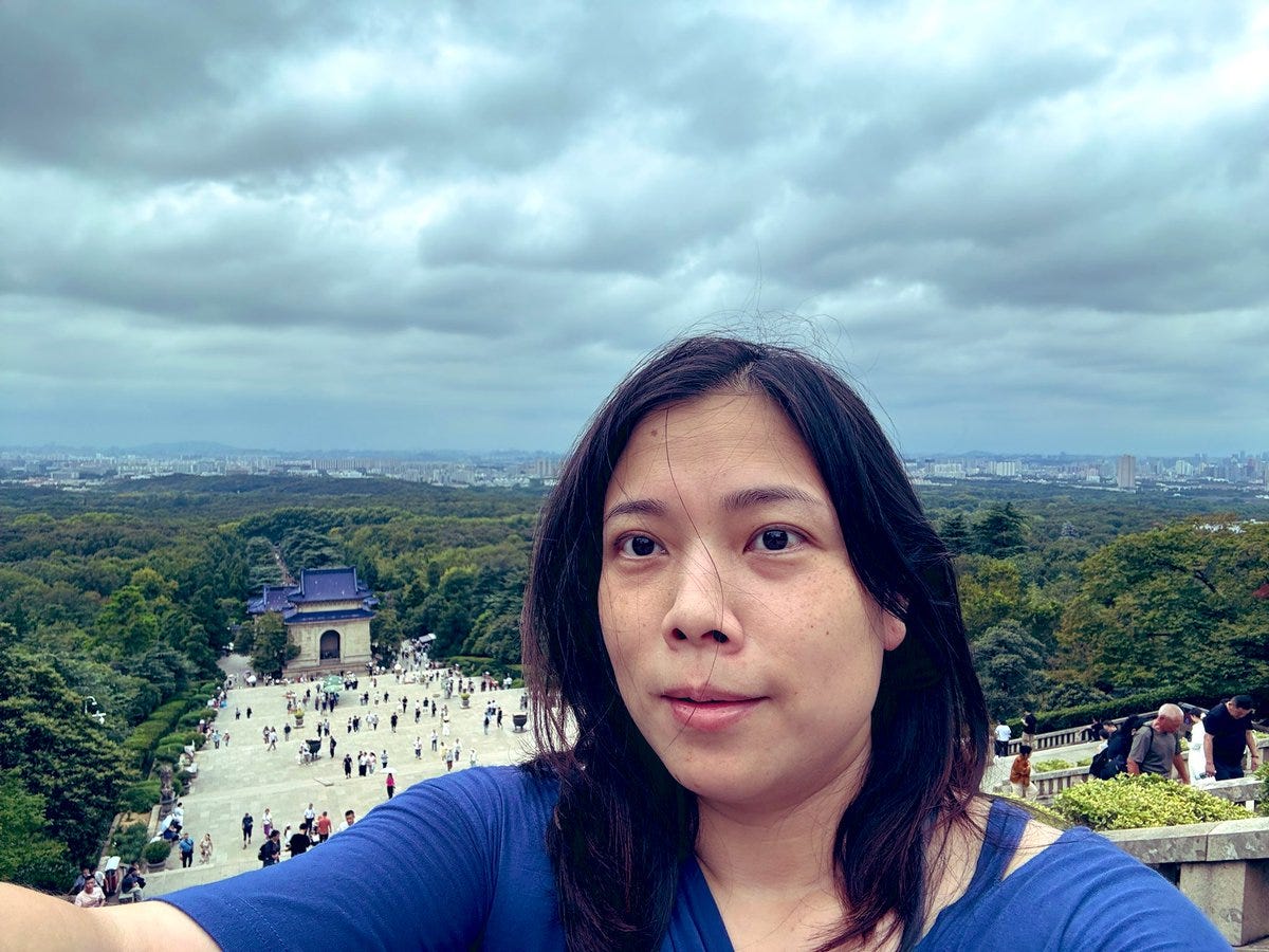A woman with long dark hair wearing a blue shirt takes a selfie, with a scenic view in the background. The background shows a large open plaza with many people, a blue and white building with the 青天白日滿地紅 KMT motif on its ceiling, and lush green hills extending to a distant city skyline under a cloudy sky.