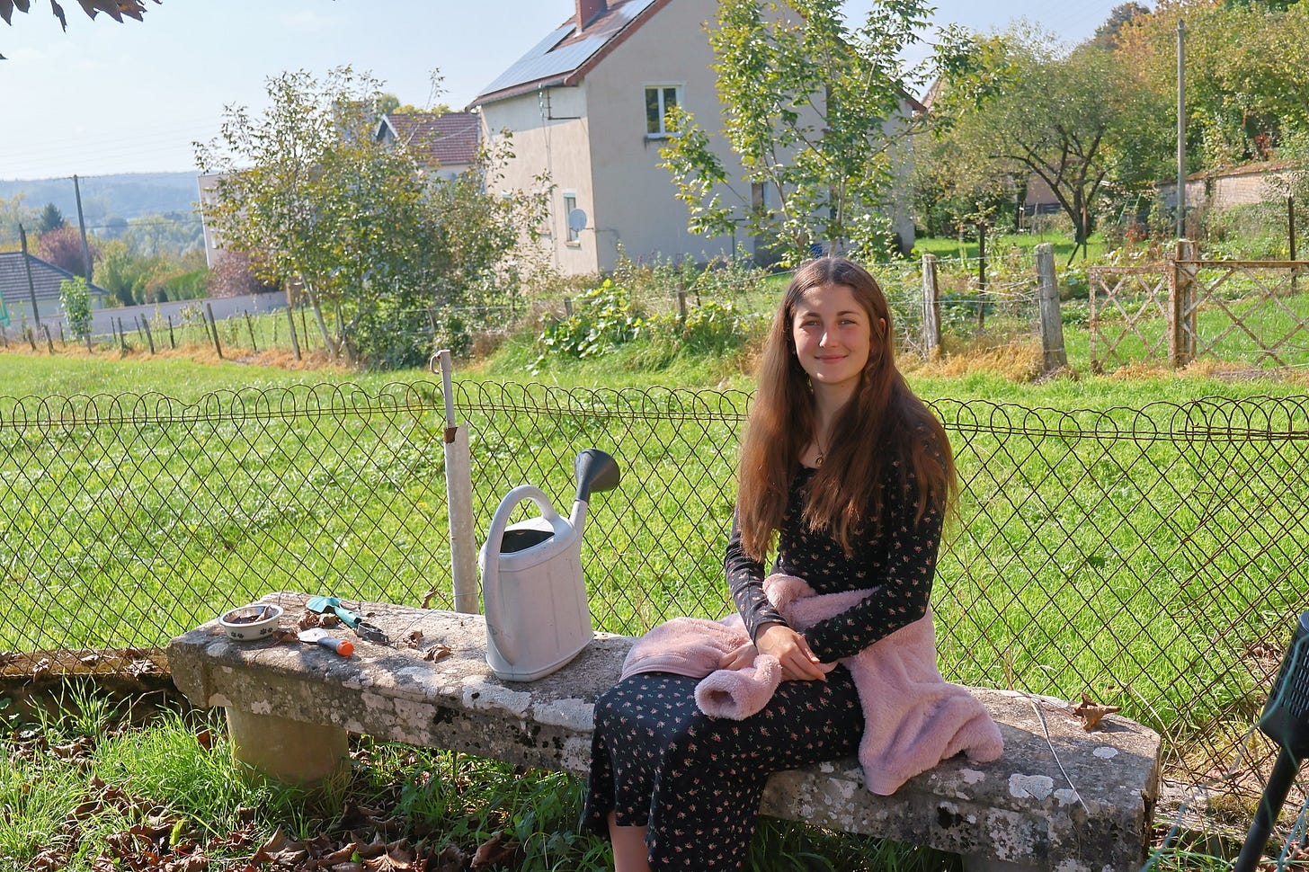 Jeune femme assise sur un banc de pierre dans un jardin rural, sous le soleil d’automne, un arrosoir à ses côtés, symbole de transmission et de mémoire vivante. Jeune femme assise sur un banc de pierre dans un jardin rural, sous le soleil d’automne, un arrosoir à ses côtés, symbole de transmission et de mémoire vivante.