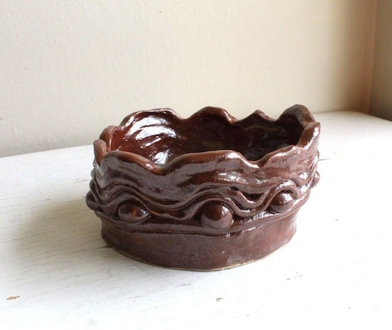 May include: A handcrafted, dark brown ceramic bowl. The bowl features a wavy, textured rim with small, decorative spheres along the edge.  The piece is a unique, handmade pottery item, ideal for decorative or functional use. The rich, dark brown glaze gives it a rustic, earthy appeal. This handmade pottery bowl is perfect for adding a touch of handcrafted charm to any home decor.