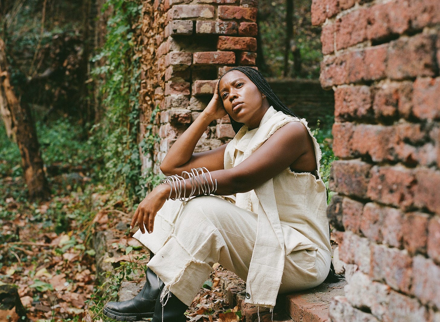 Image Description: I am wearing long black braids, an unbleached cotton jumpsuit canvas jumpsuit I made from drop cloth, black rubber boots and an armature wire arm cuff sculpted around my arm while gazing away from the camera and sitting inside an opening of a decaying brick wall being reclaimed by the forest. Photography: Chukwudumebi Ezefili Image Description: I am wearing long black braids, an unbleached cotton jumpsuit canvas jumpsuit I made from drop cloth, black rubber boots and an armature wire arm cuff sculpted around my arm while gazing away from the camera and sitting inside an opening of a decaying brick wall being reclaimed by the forest. Photography: Chukwudumebi Ezefili
