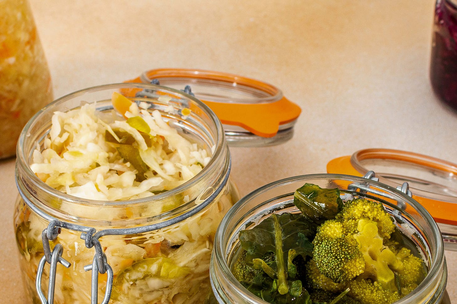 Two open clamp-lid jars with fermented vegetables rest on a kitchen countertop: on the left, sauerkraut with spices; on the right, broccoli and leafy greens submerged in brine. The orange rubber sealing rings lie next to the jars.