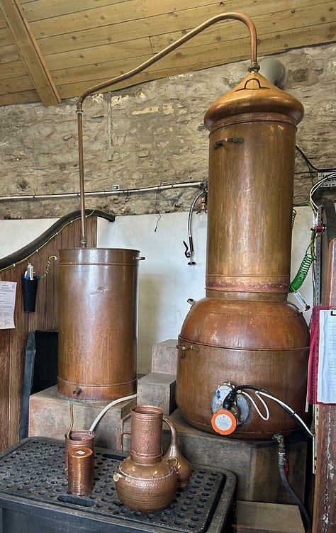A box of flowers and leaves, a large copper still, shelves of bottles for sale