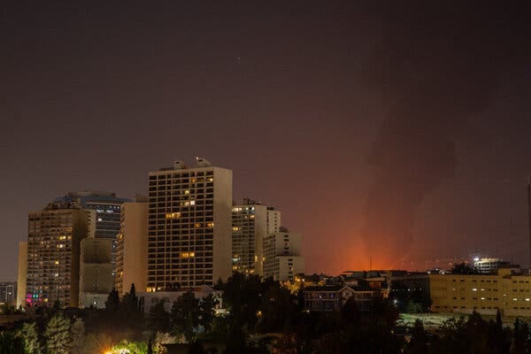Large buildings in Tehran, at night; the sky is lit up behind them from an airstrike.