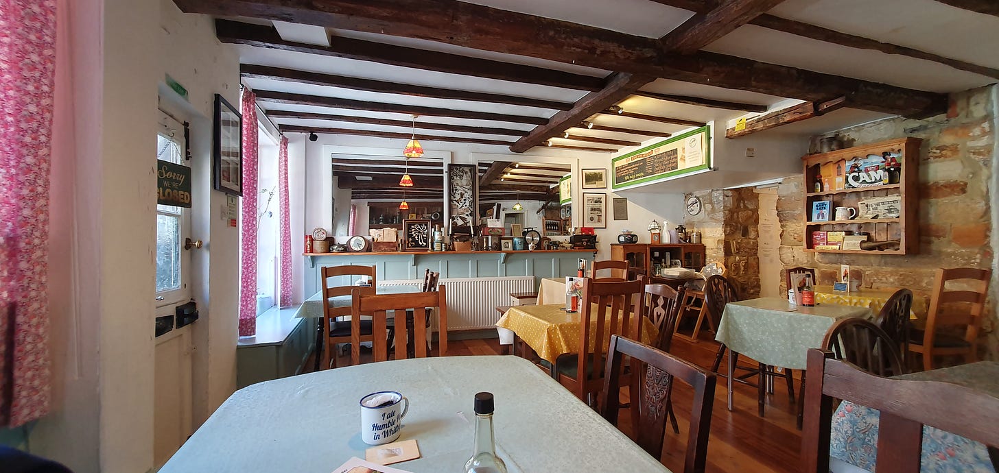 An inside photo of the Humble Pie and Mash, mismatched wooden tables are scattered throughout the room, wooden chairs and 1940s memorabilia lines the walls. Music from between the wars and the 1940s can be heard. Click on this image and you can listen to the music via a YouTube upload of this scene.