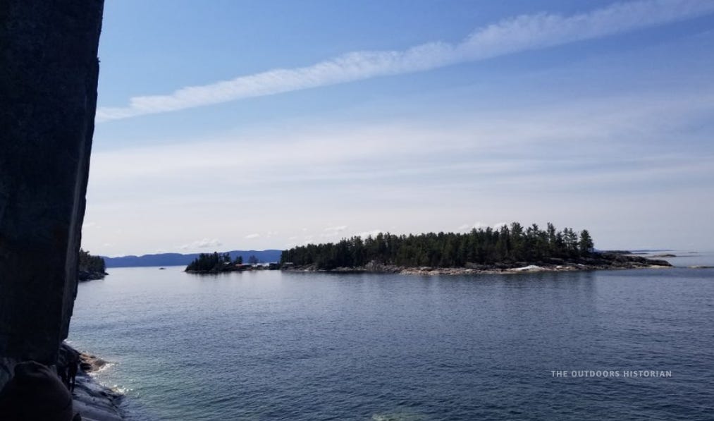 The sheer rock wall at Agawa Rock featuring red ochre pictographs, with the Agawa Islands visible in the distance on a sunny May day with lingering snow.