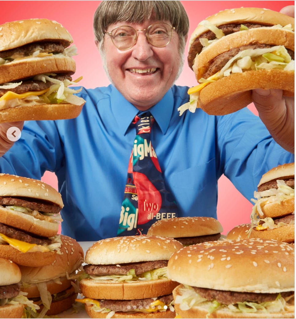 Donald Gorske, holds a Big Mac in each hand with many more ready to be eaten on the table. He has a colorful, wide tie with the words "BigMac" and "Two all-BEEF Patties"