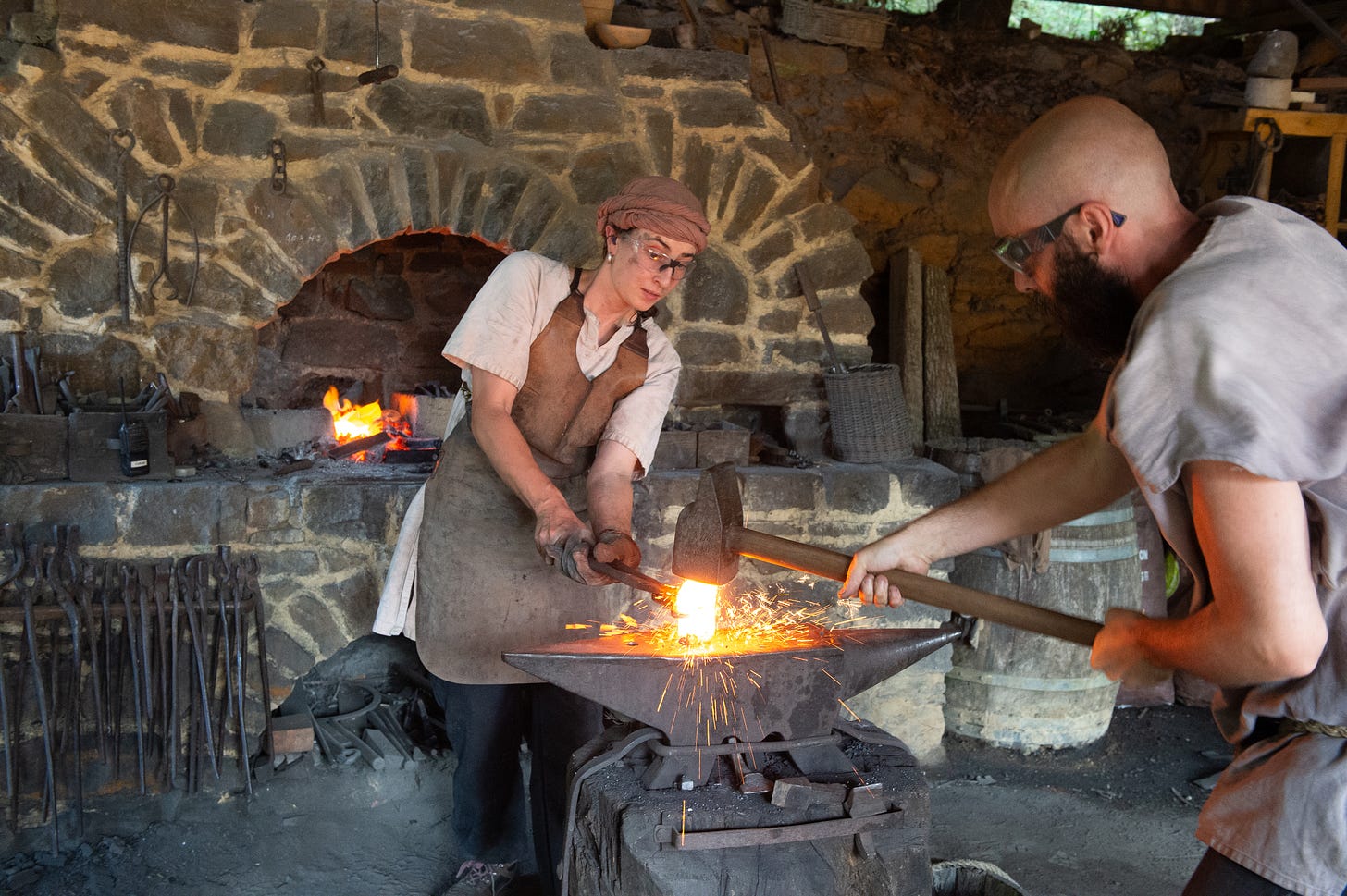 Two blacksmiths forge heated metal on an anvil, with sparks flying inside a traditional stone forge workshop. Two blacksmiths forge heated metal on an anvil, with sparks flying inside a traditional stone forge workshop.