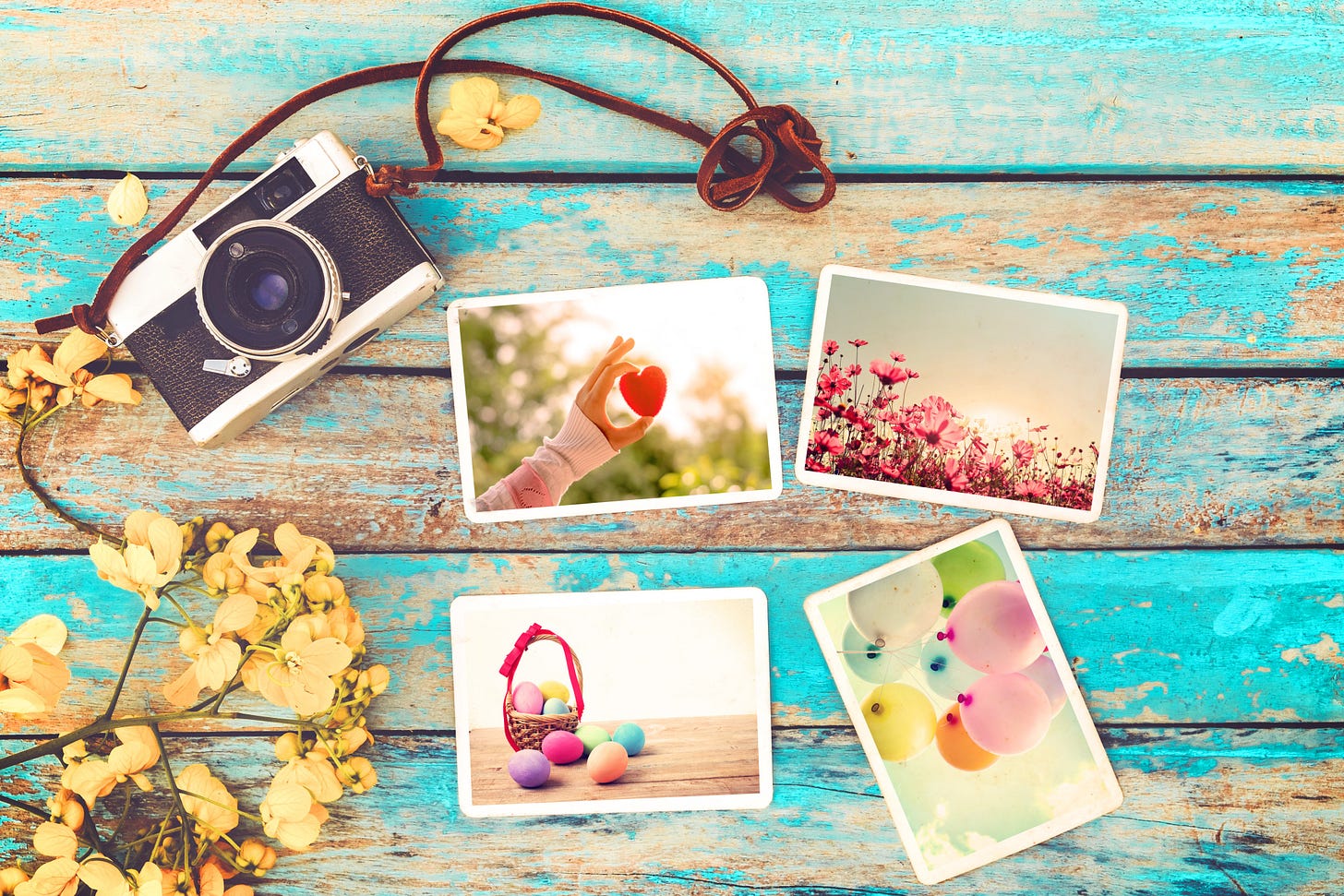 Weathered, blue wooden background. Over it there’s a vintage camera, yellow flowers and 4 photographs: a hand holding a small red heart against the sky, pink flowers in the sunset, a small wicker basket filled and surrounded by colorful Easter eggs, and a cluster of pastel colored balloons.