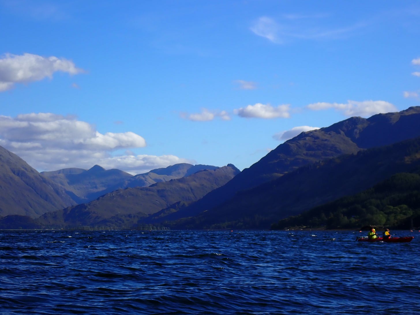 Deep blue water with a small boat holding two people to the right, with layers of mountains and blue sky behind