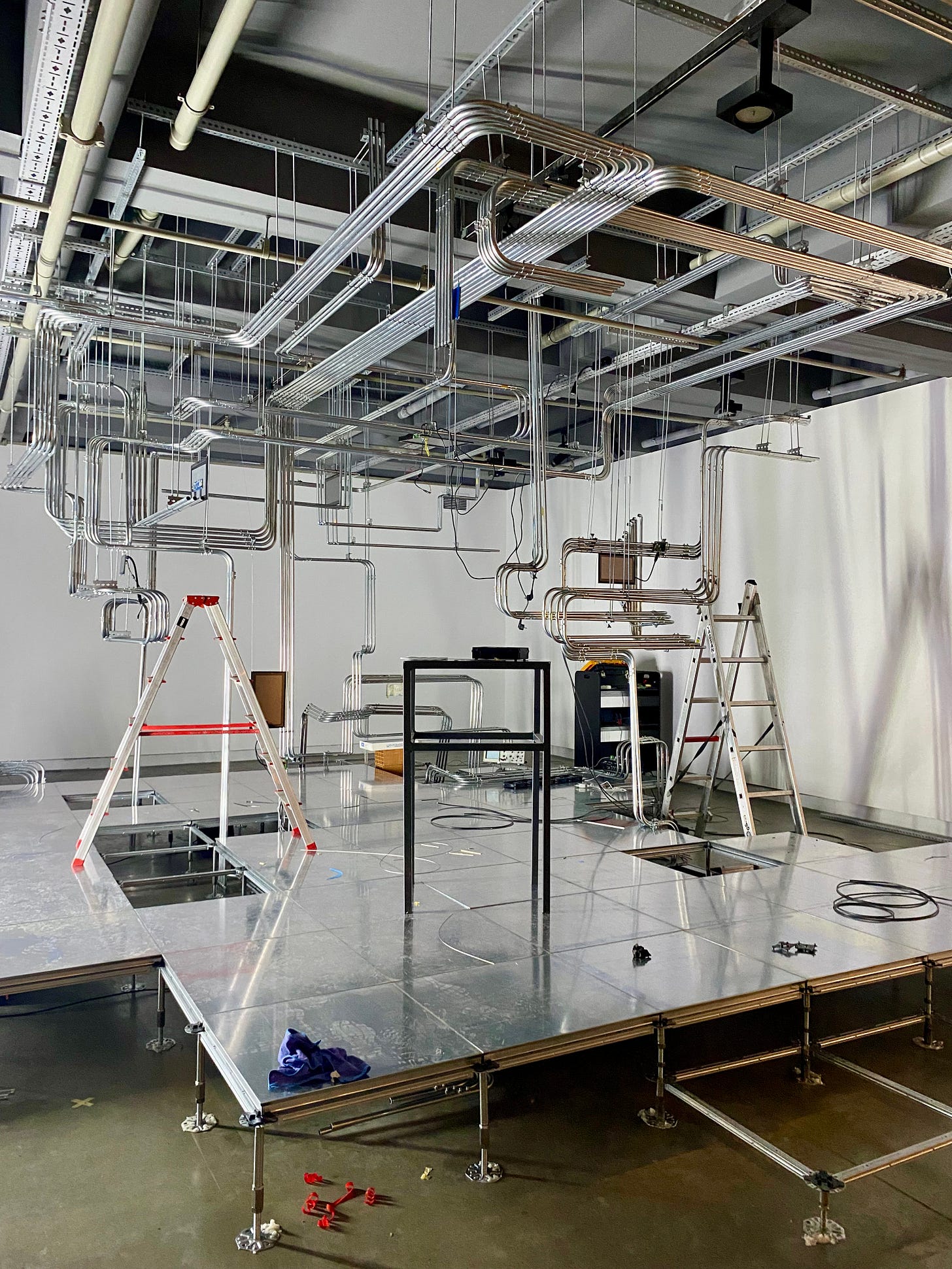 A gallery space with spare interior fittings and an art installation under construction showing a raised platform with a series of steel pipes, running in fixed track formation with occasional bends, that range from the ceiling to the floor in the middle of which are a number of small flat screens with graphical displays.