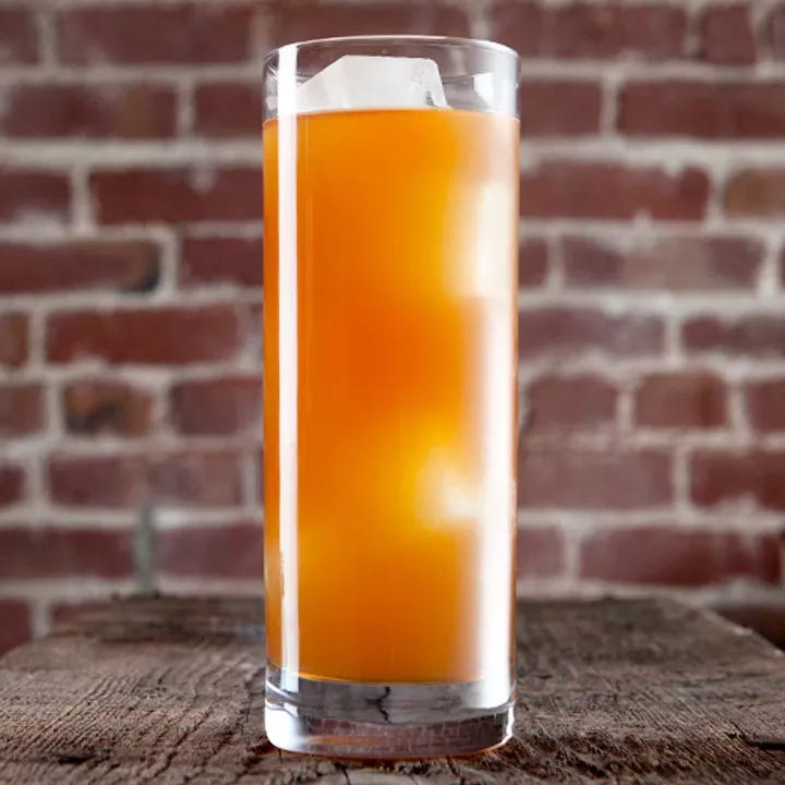 A tall Collins glass sits on a rustic wooden table, a brick wall in the background. The drink within is bright orange and cloudy, with a few large ice cubes floating in it. A tall Collins glass sits on a rustic wooden table, a brick wall in the background. The drink within is bright orange and cloudy, with a few large ice cubes floating in it.