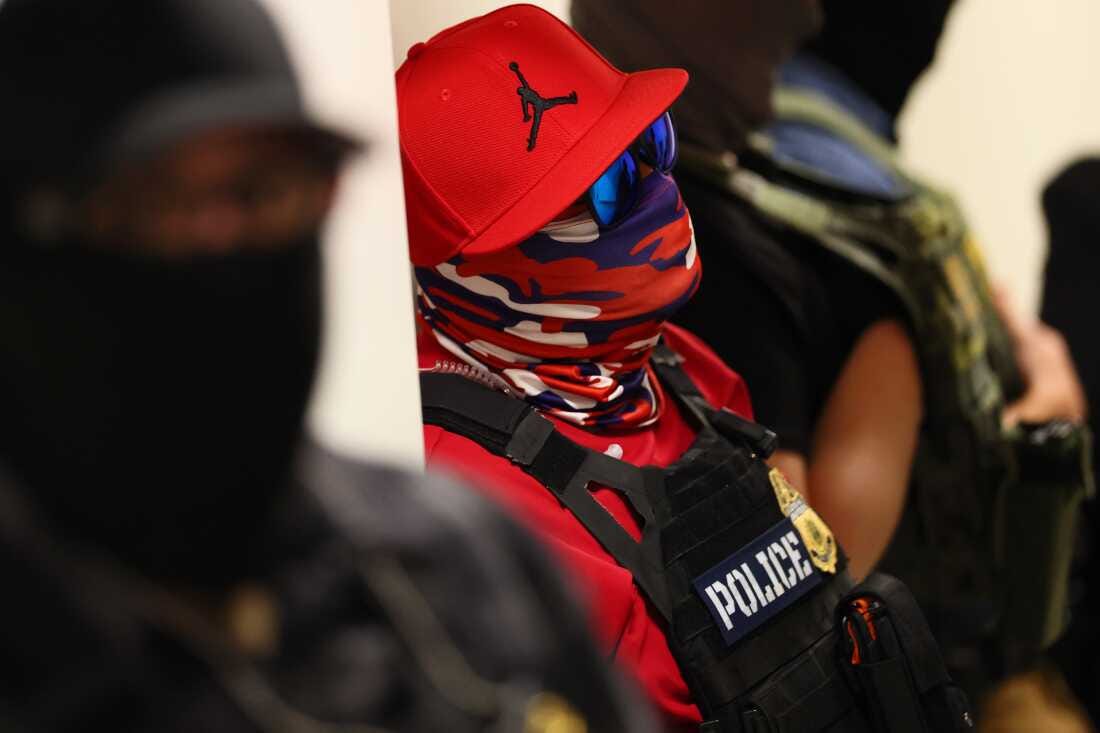 Federal agents wearing masks patrol the halls of the immigration court at the Ted Weiss Federal Building in New York City on July 9. The agent in the center of the photo is wearing a red baseball cap with the Air Jordan logo on it, sunglasses with blue lenses, a red and blue bandana that covers the face, a red shirt and a black vest that says "POLICE" on the front. Federal agents wearing masks patrol the halls of the immigration court at the Ted Weiss Federal Building in New York City on July 9. The agent in the center of the photo is wearing a red baseball cap with the Air Jordan logo on it, sunglasses with blue lenses, a red and blue bandana that covers the face, a red shirt and a black vest that says "POLICE" on the front.